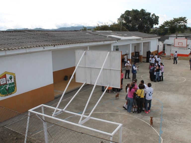 Vista desde lo alto de la Institución Educativa Alto Nubia: aulas de muros blancos alrededor de la cancha, con la comunidad reunida en el patio.