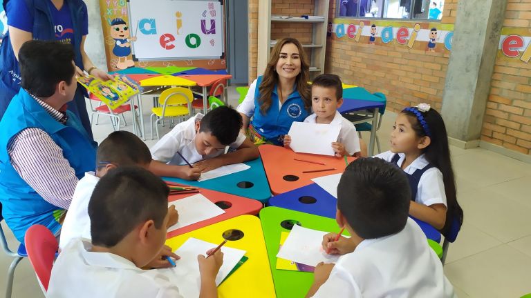 Niños dibujando en una mesa de piezas de colores, acompañados por una representante de la Fundación, frente a un tablero con las vocales.