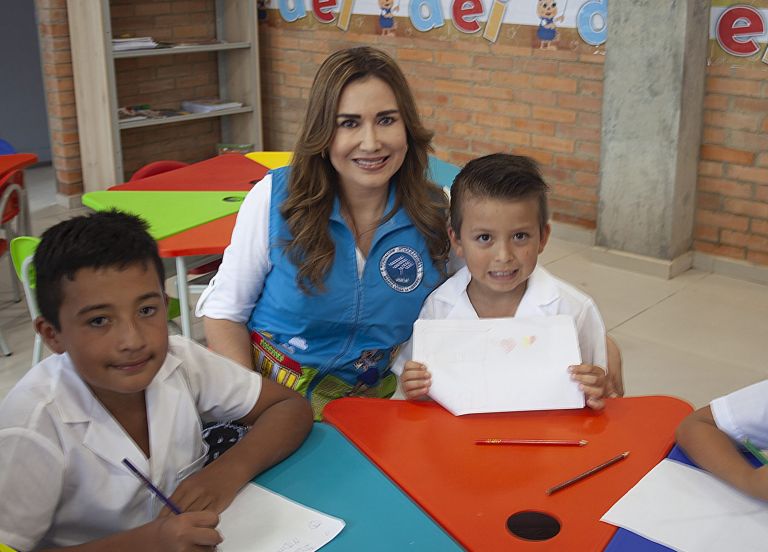 Un niño muestra su dibujo junto a un compañero y a una representante de la Fundación, sentados en una de las mesas de colores.