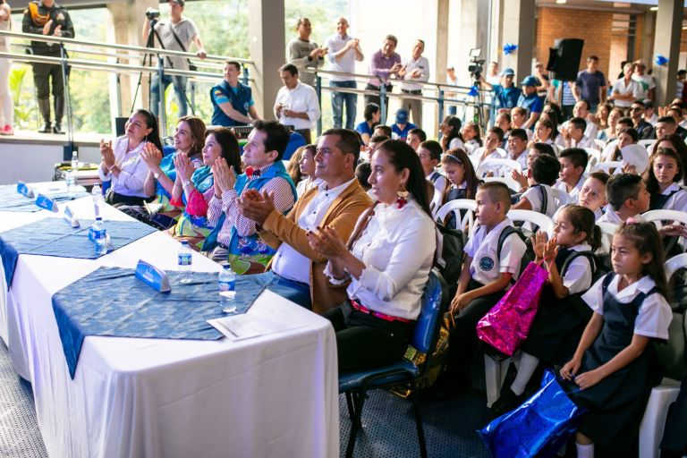 La mesa principal y los estudiantes uniformados aplauden durante la ceremonia, con sus bolsas de regalo en las manos.