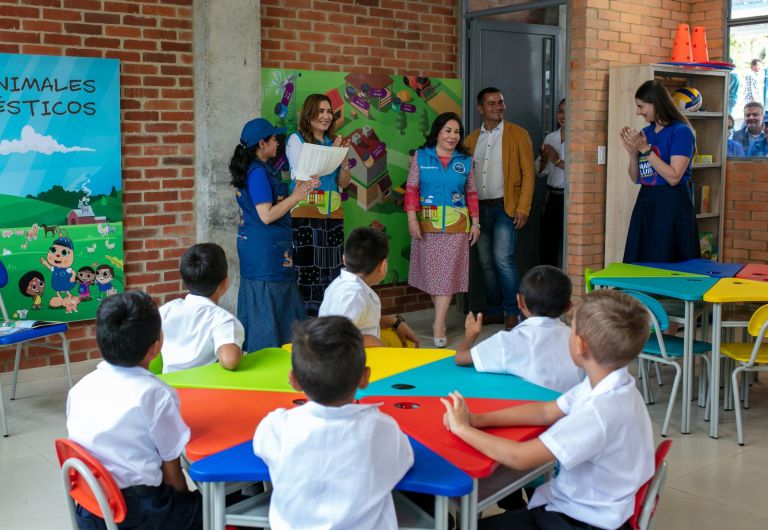 La Dra. María Luisa de Moreno y el equipo de la Fundación entran al aula de preescolar mientras los niños, sentados en las mesas de colores, los reciben.