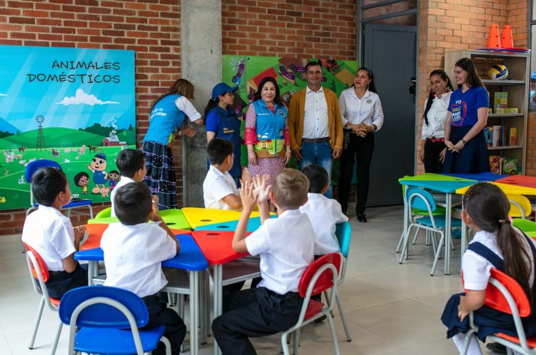 Los niños saludan con la mano levantada a la Dra. María Luisa de Moreno, al alcalde y a las voluntarias, de pie junto a los murales del aula.