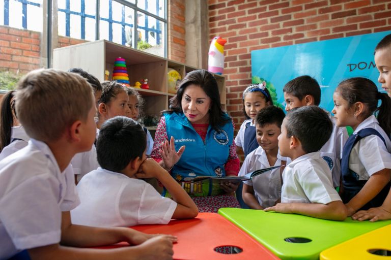 Rodeada por los niños alrededor de las mesas de colores, la Dra. María Luisa de Moreno les cuenta una historia con el libro abierto.