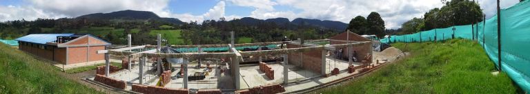 Panorámica de la obra en curso: columnas de concreto y muros de ladrillo a media altura sobre la placa, con el primer bloque ya techado al fondo y las montañas de la sabana detrás.