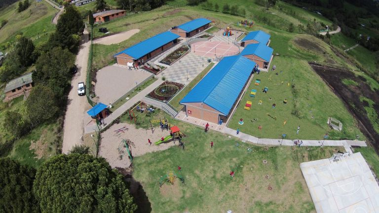 Vista aérea de la sede terminada: cuatro bloques de aulas con cubierta azul alrededor de una plazoleta ajedrezada, con parque infantil, jardines y cancha.