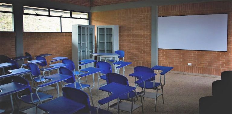 Un aula terminada y dotada: pupitres azules nuevos, tablero blanco, armarios metálicos y ventanales que dan al campo.
