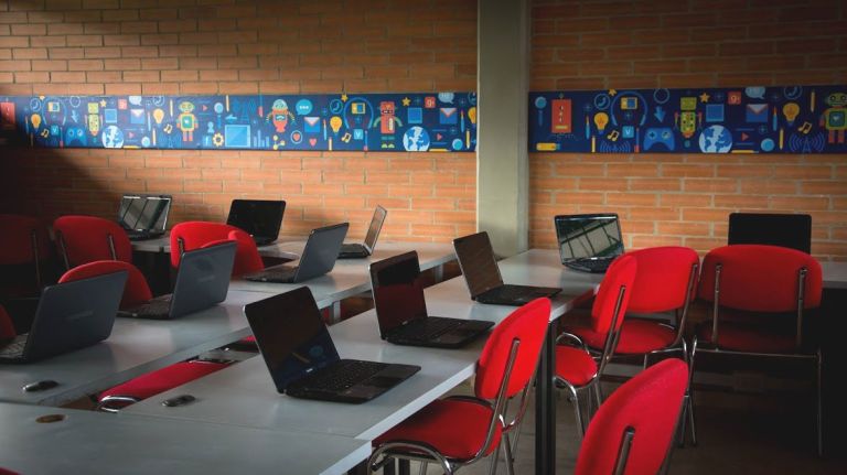 La sala de sistemas: portátiles abiertos sobre mesas blancas con sillas rojas, frente a una cenefa decorada con íconos de tecnología.