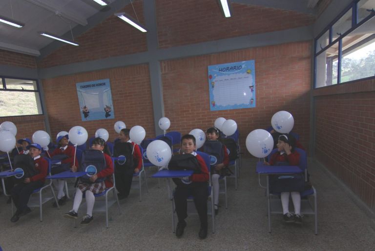 Los estudiantes estrenando un aula el día de la entrega, sentados en sus pupitres con globos blancos y sus maletas nuevas.