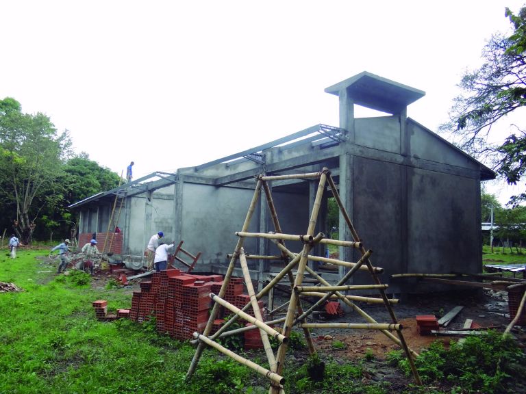 La construcción en curso: los muros y la estructura de concreto levantados, con andamios de guadua y obreros trabajando en la cubierta.