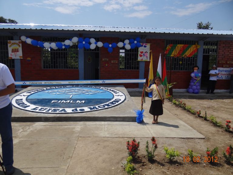 La escuela el día de la entrega, adornada con globos azules y blancos y carteles de bienvenida, con el sello de la Fundación fundido en la plazoleta y una estudiante con la banda tricolor.