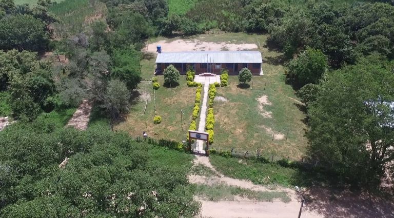 Vista aérea de la escuela terminada: el bloque de aulas con cubierta metálica, el sendero de acceso bordeado de arbustos y el portal de entrada, rodeado de monte.