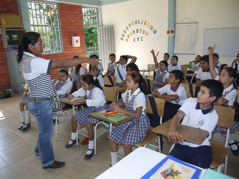 Una clase en marcha: el docente de pie frente al grupo y los estudiantes con uniforme a cuadros levantando la mano desde sus pupitres.