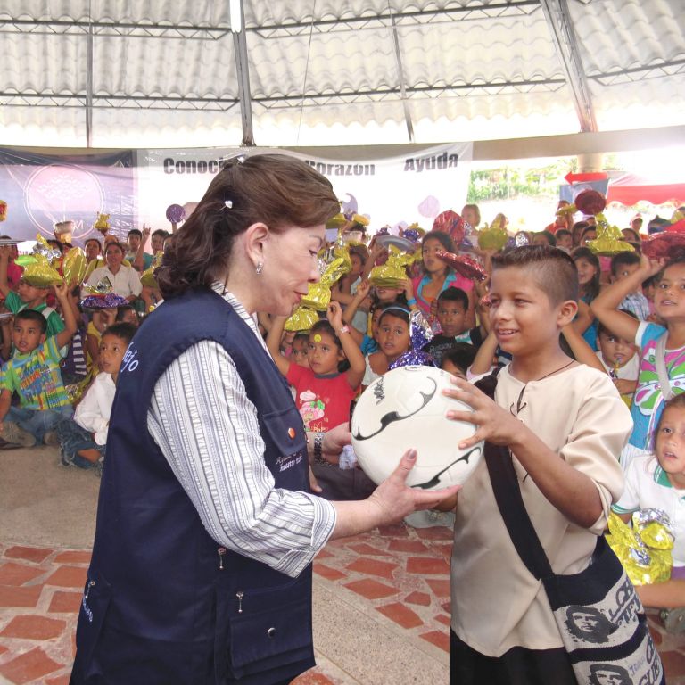 La Dra. María Luisa de Moreno entrega un balón a un estudiante, frente a los demás niños que levantan sus regalos.