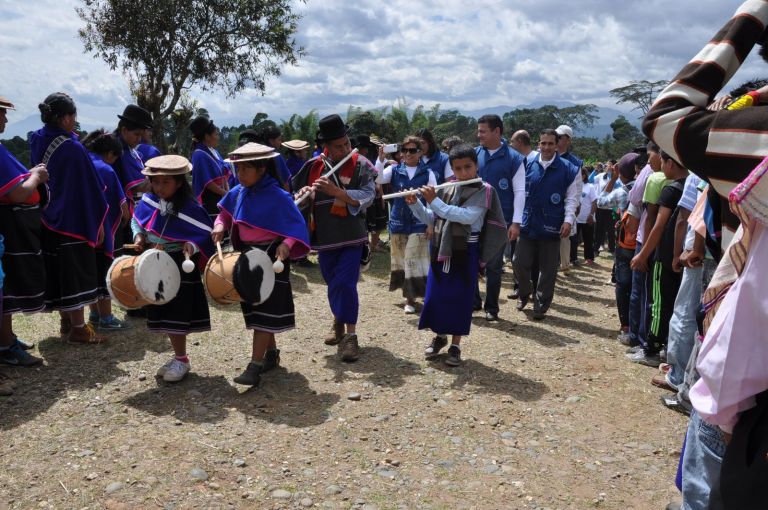 Dos niñas misak con sombrero tejido y ruana azul tocan tambores al frente del desfile de llegada, seguidas por un niño que toca la flauta y por el equipo de la Fundación, entre dos filas de la comunidad.