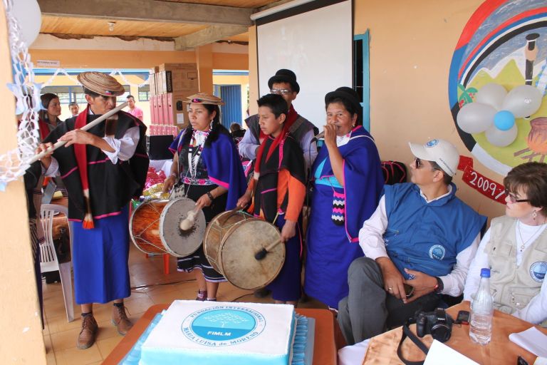 Músicos misak tocan tambores y flauta y una mujer canta en el corredor de la escuela; al fondo, las cajas de los computadores entregados y, delante, una torta con el sello de la Fundación.