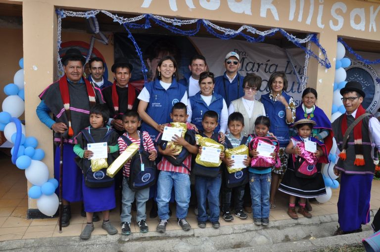Niñas y niños de la escuela con sus morrales nuevos, regalos y certificados, junto a autoridades misak y al equipo de la Fundación, frente a un salón decorado con globos azules y blancos.