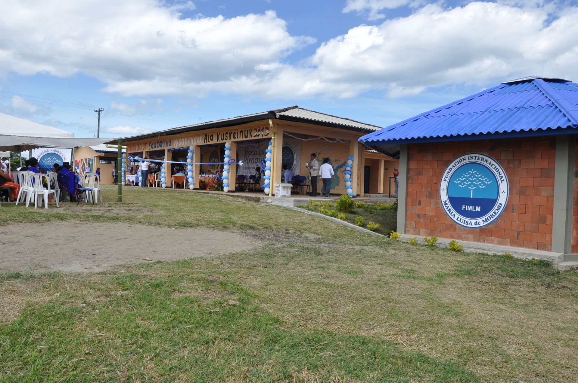 La escuela el día de la inauguración: un corredor de aulas color durazno con el letrero Institución Educativa Misak Misak Ala Kusreinuk 2013, decorado con globos azules y blancos, y a la derecha el nuevo salón de ladrillo y techo azul con el sello de la Fundación.