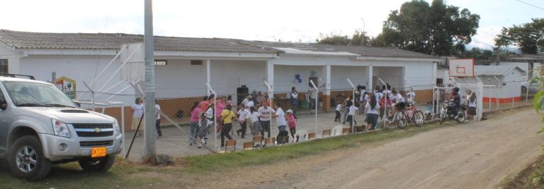 La escuela vista desde el camino de tierra: un bloque de aulas blancas con zócalo naranja y corredor de columnas, y la cancha cercada donde niños y adultos juegan en ronda el día de la entrega.
