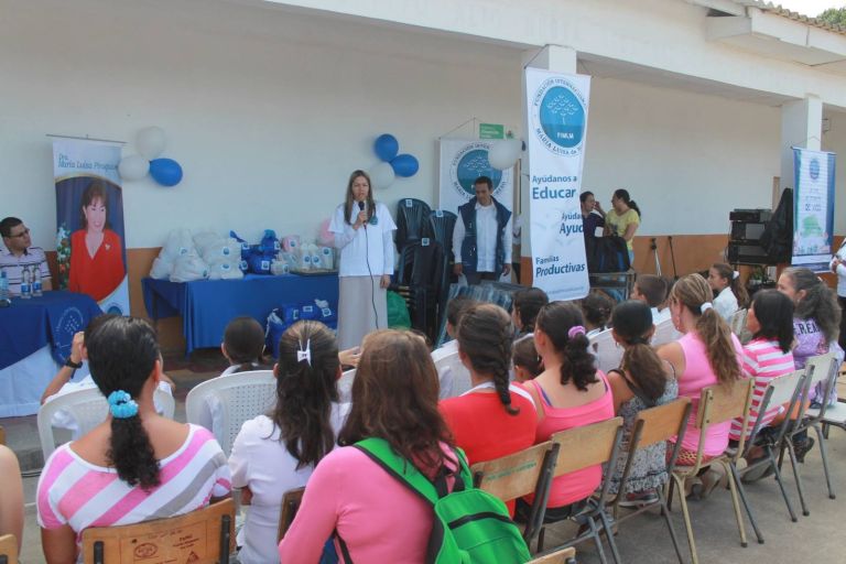 Una representante de la Fundación habla al micrófono ante madres y estudiantes sentados en el corredor de la escuela, junto a los paquetes de kits y a los pendones Ayúdanos a Educar.