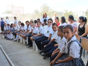 Estudiantes de uniforme sentados en fila en la cancha, con sus madres detrás, durante el acto de entrega.