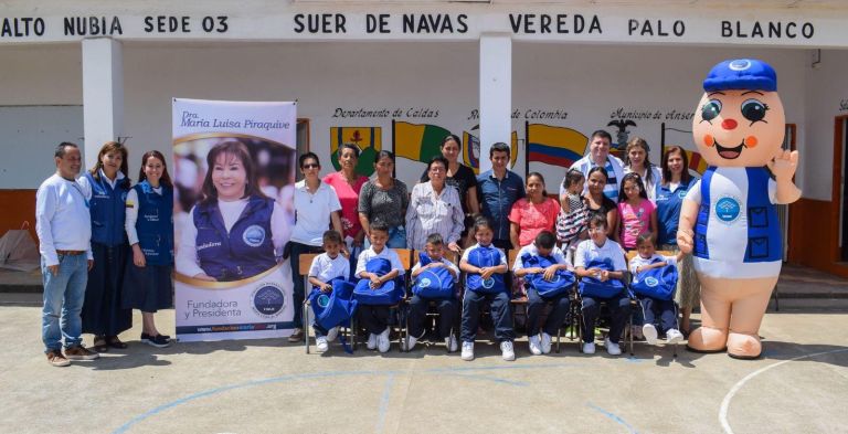 Años después, en una entrega de Mi Nueva Familia: niños de uniforme con morrales azules, sus familias y voluntarios de la Fundación frente al letrero Alto Nubia sede 03 Suer de Navas, vereda Palo Blanco.