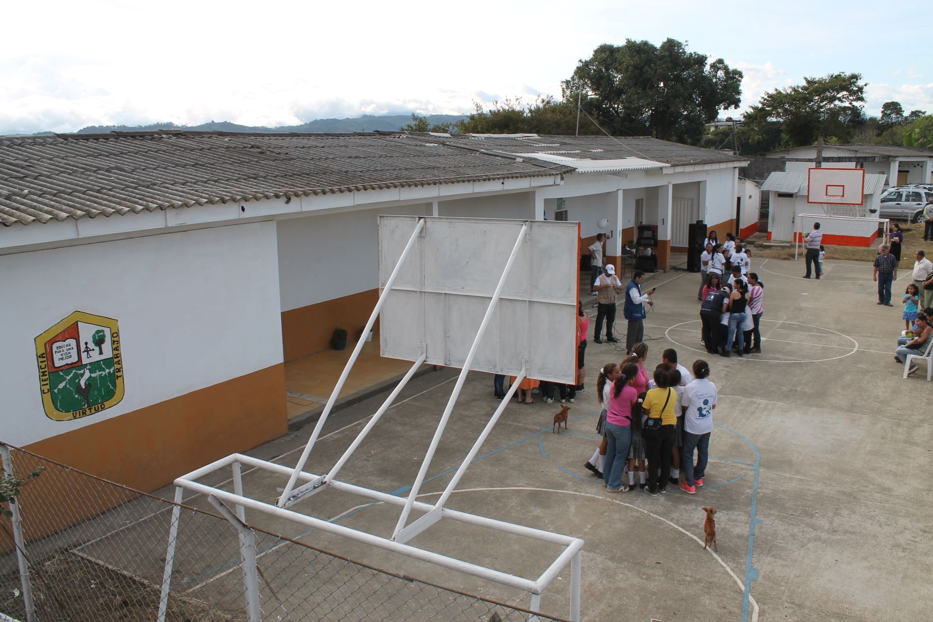 La escuela el día de la entrega, vista desde lo alto: aulas blancas con zócalo naranja y el escudo «Ciencia, virtud, trabajo» pintado en el muro, y la cancha donde estudiantes, familias y voluntarios de la Fundación se reúnen.