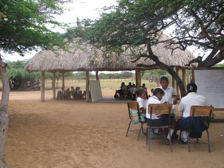 Antes de la nueva sede: estudiantes con uniforme estudian en pupitres sobre la arena, unos bajo una enramada de palma y otros a la sombra de los árboles, frente a un tablero.
