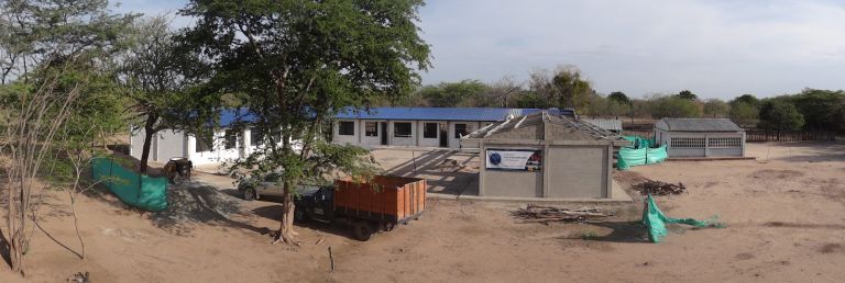 La obra en marcha: el bloque de aulas ya techado de azul y un salón nuevo en construcción, con un camión de materiales y polisombra verde alrededor.