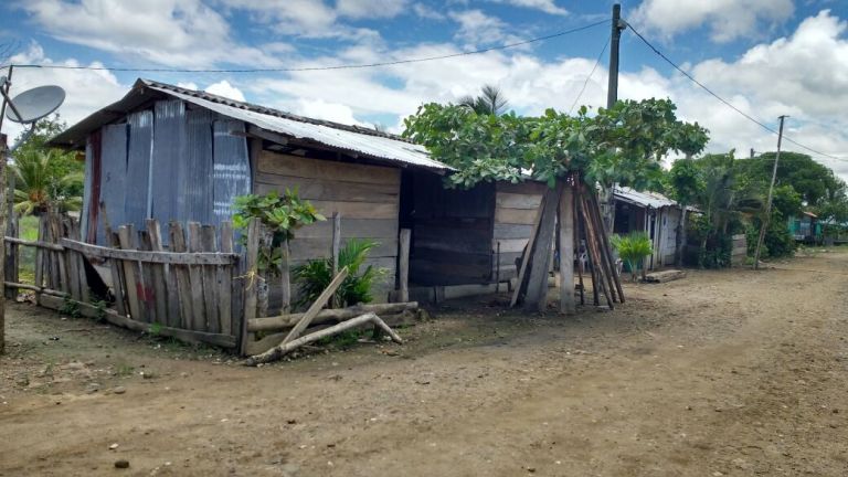 La vereda antes de la escuela: casas de tablas y láminas de zinc a lado y lado de un camino de tierra, con cercas de madera y palmeras.