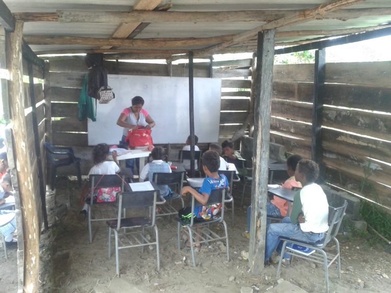 Una clase en el salón de tablas que prestó un vecino: la docente frente al tablero y los niños en pupitres metálicos, sobre piso de tierra y bajo un techo de zinc sostenido con troncos.