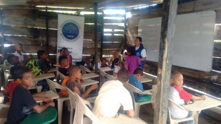 Niños de distintas edades compartiendo el mismo salón de tablas desiguales, con sillas plásticas, mientras una voluntaria de la Fundación les habla junto al pendón de Ayúdanos a Educar.