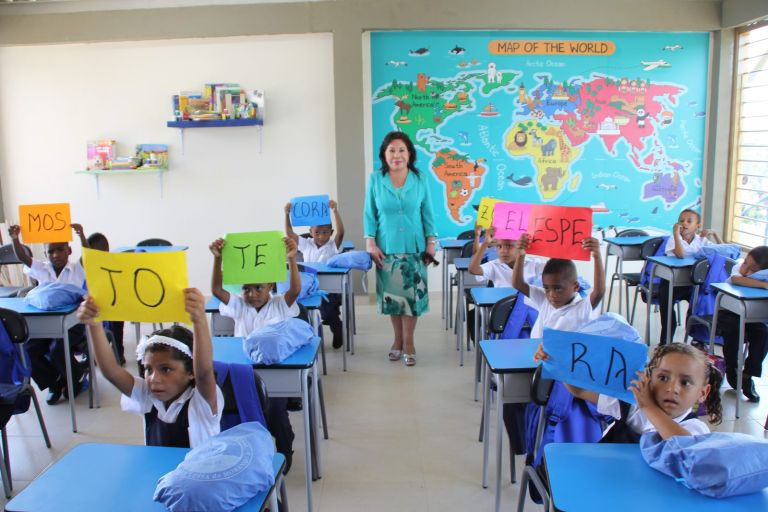 En un aula nueva con un gran mapa del mundo en la pared, los niños levantan carteles de colores con sílabas mientras la Dra. María Luisa de Moreno los acompaña.