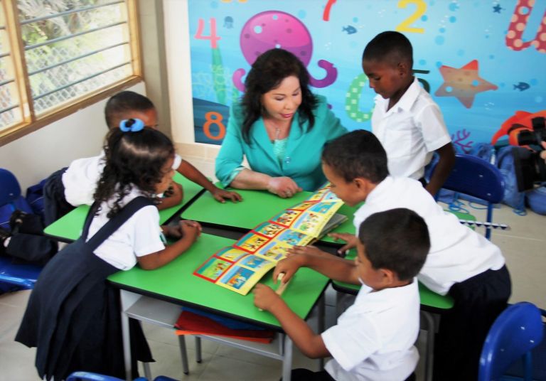 La Dra. María Luisa de Moreno lee un libro ilustrado con un grupo de niños alrededor de una mesa verde, frente a un mural de números y animales marinos.