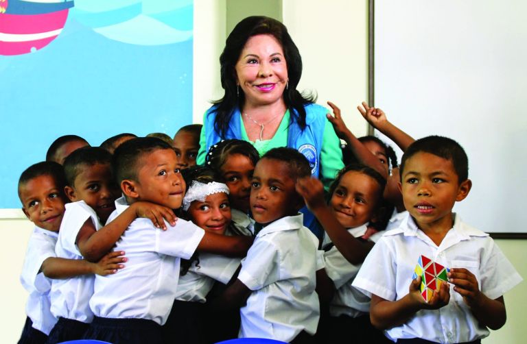 Un grupo de niños abraza a la Dra. María Luisa de Moreno en el aula; uno de ellos sostiene un juguete de colores.