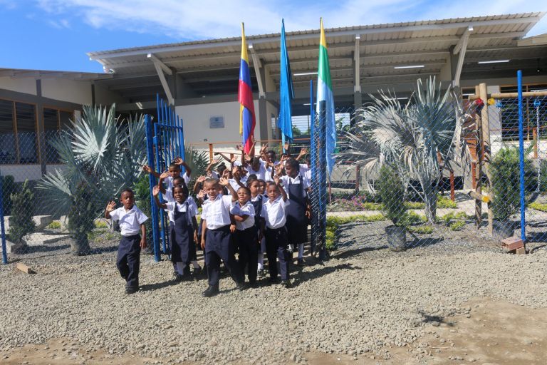 Los estudiantes salen por la reja de la nueva escuela saludando con las manos en alto, frente a las banderas y los jardines de palmas.