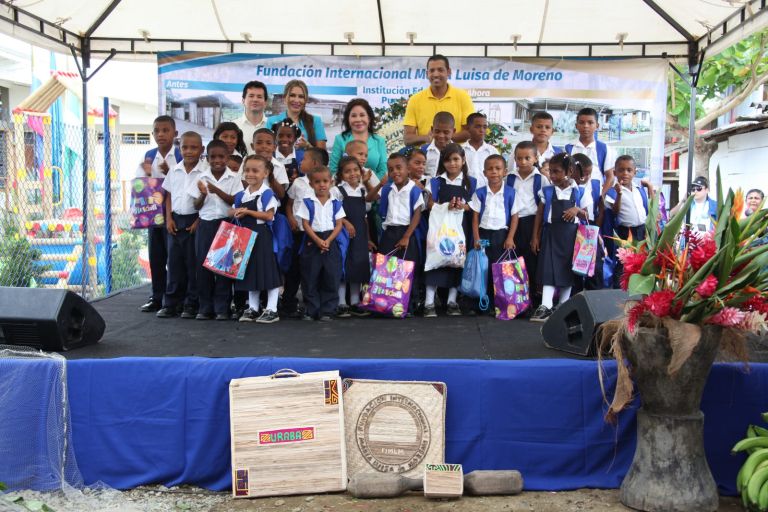 Los niños con uniforme y maletas nuevas posan en la tarima con sus regalos, delante de un pendón que muestra el antes y el después de la escuela.