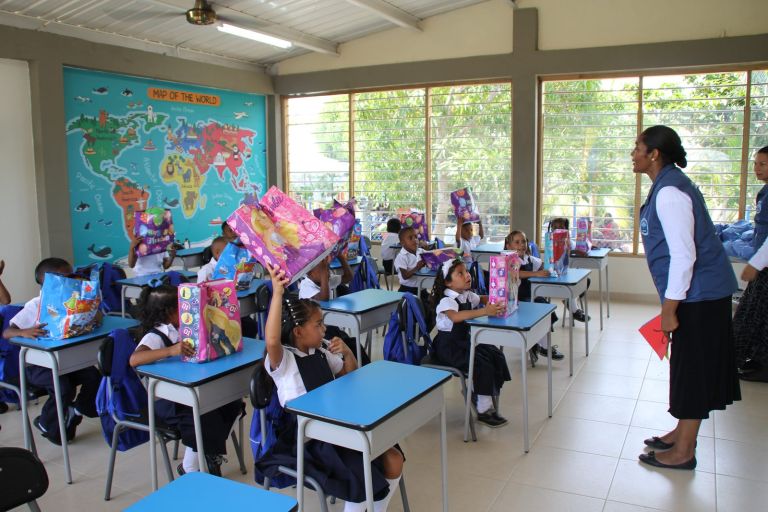 En un aula nueva con un gran mapa del mundo, los niños levantan las bolsas de regalo mientras una voluntaria les habla.