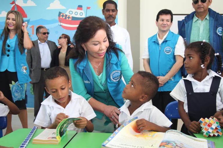 La Dra. María Luisa de Moreno conversa con un niño que hojea un libro en su pupitre verde, rodeada por otros estudiantes y por el equipo de la Fundación.