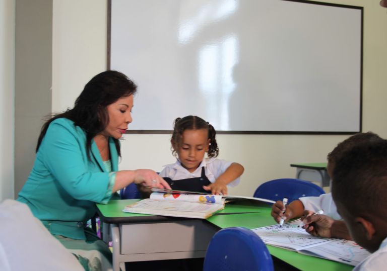 La Dra. María Luisa de Moreno lee junto a una niña en el aula nueva, mientras otros compañeros dibujan en la misma mesa.