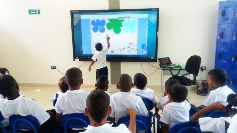 Un niño resuelve un ejercicio en el tablero digital del aula mientras sus compañeros lo observan sentados.