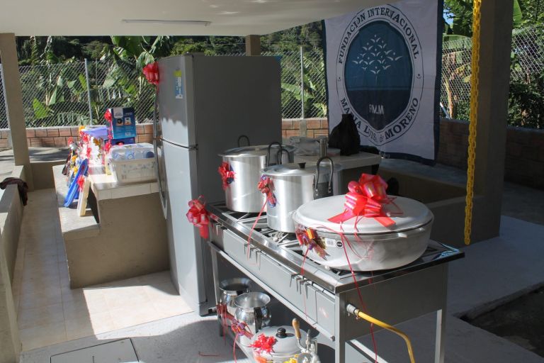 La cocina de la escuela con su dotación nueva, adornada con moños rojos: nevera, estufa industrial con ollas grandes y, al fondo, el pendón de la Fundación.
