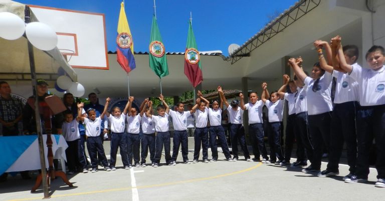 Los estudiantes, con uniforme blanco y azul nuevo, se toman de las manos y levantan los brazos en la cancha, frente a las banderas de Colombia, Risaralda y Belén de Umbría.