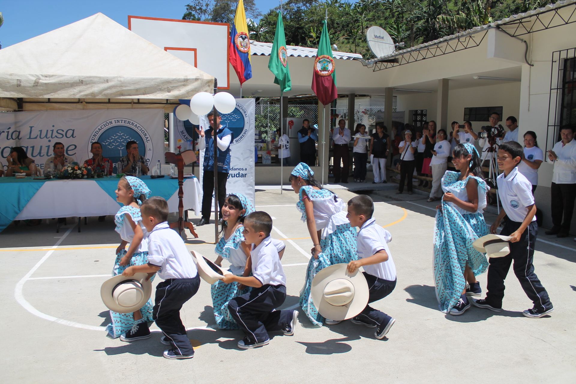 Niñas y niños de la escuela bailan con trajes típicos y sombreros en la cancha renovada, frente a la sede pintada de blanco y a las banderas de Colombia, Risaralda y Belén de Umbría, mientras los invitados aplauden.