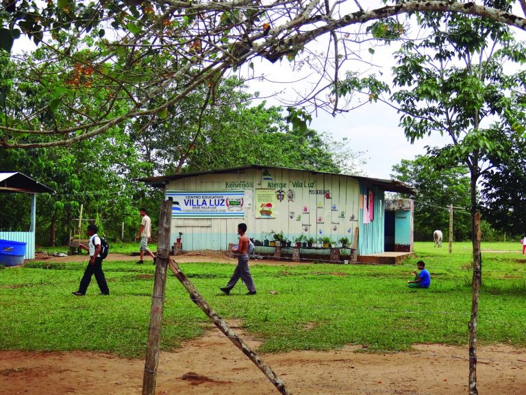 La antigua sede: una construcción de tablas pintada de blanco con el letrero «Centro Educativo Villa Luz» y la leyenda «Bienvenidos Albergue Villa Luz», rodeada de potrero, con estudiantes caminando frente a ella.