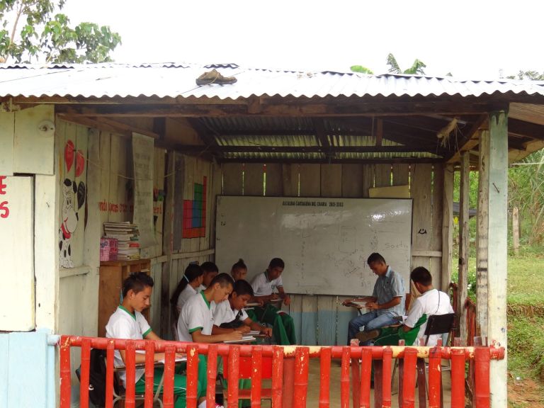 Una clase en la sede antigua: estudiantes con uniforme verde y blanco escriben en sus pupitres bajo un techo de zinc sostenido con vigas de madera, frente a un tablero.