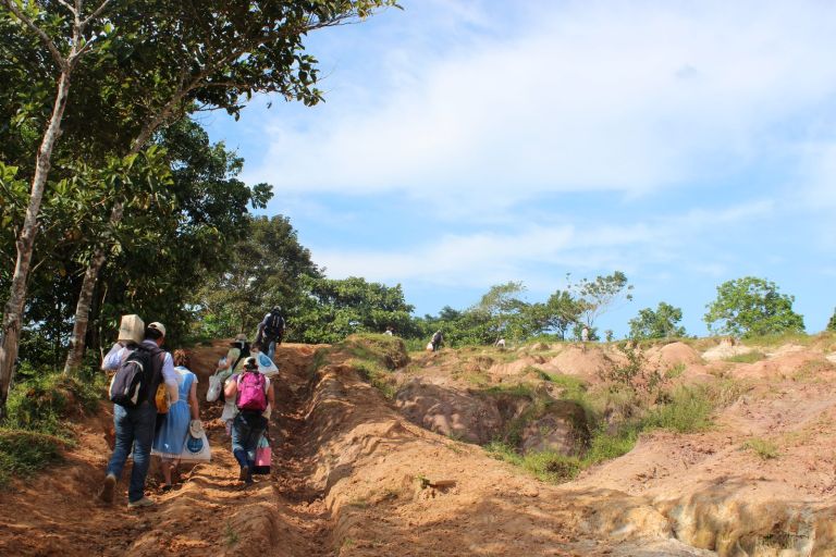 Voluntarios suben a pie por un camino de tierra erosionado, cargando cajas y bolsas, en el último tramo hacia la escuela.