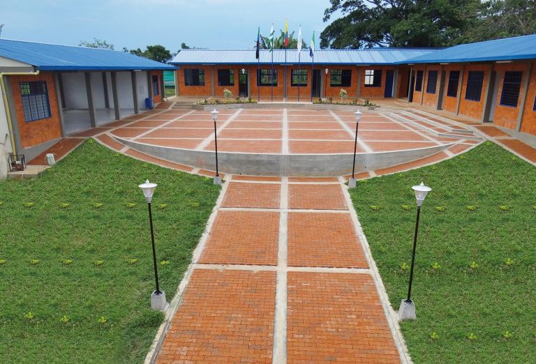 La sede terminada: bloques de aulas en ladrillo con techos azules alrededor de una plazoleta de adoquín, con faroles, banderas y prados.