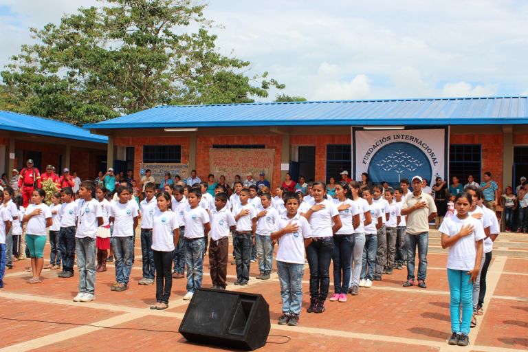 Los estudiantes cantan el himno con la mano en el pecho en la plazoleta nueva, frente a carteles de agradecimiento a la Fundación hechos por la comunidad.