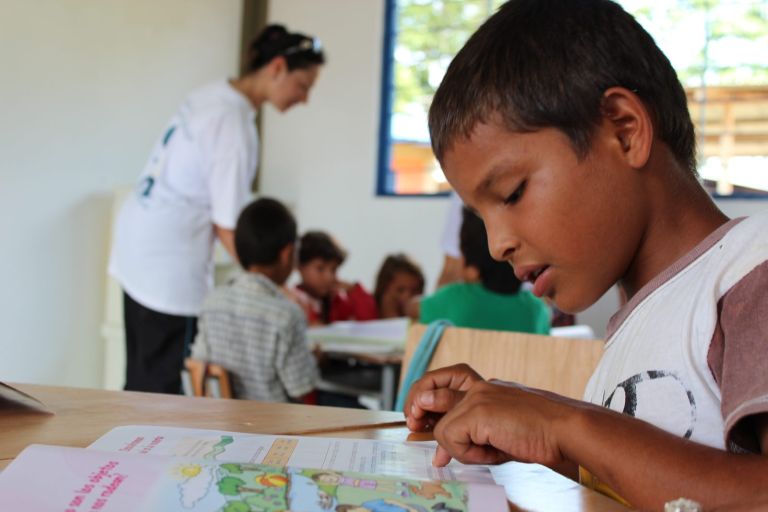 Un niño sigue con el dedo la lectura de su libro en el aula nueva, mientras al fondo una voluntaria ayuda a otros compañeros.