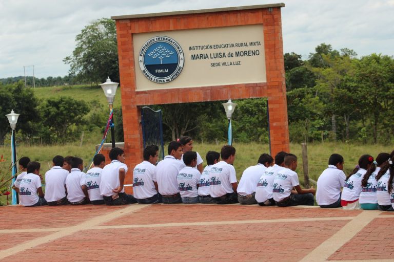 Los estudiantes, sentados de espaldas en el borde de la plazoleta, esperan frente al portal con el nombre de la institución y el escudo de la Fundación.